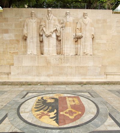 Sculptures of the four great figures of the geneva protestant movement : Guillaume Farel (1489 - 1565), Jean Calvin (1509-1564), Theodore de Beze (1513-1605) and John Knox (1513-1572) on the reformation wall in Bastions park in front of Geneva flag on the ground, Geneva, Switzerland. The construction of this wall started in 1909. The motto of the Reformation and Geneva is written behind these statues : "Post Tenebras Lux.".